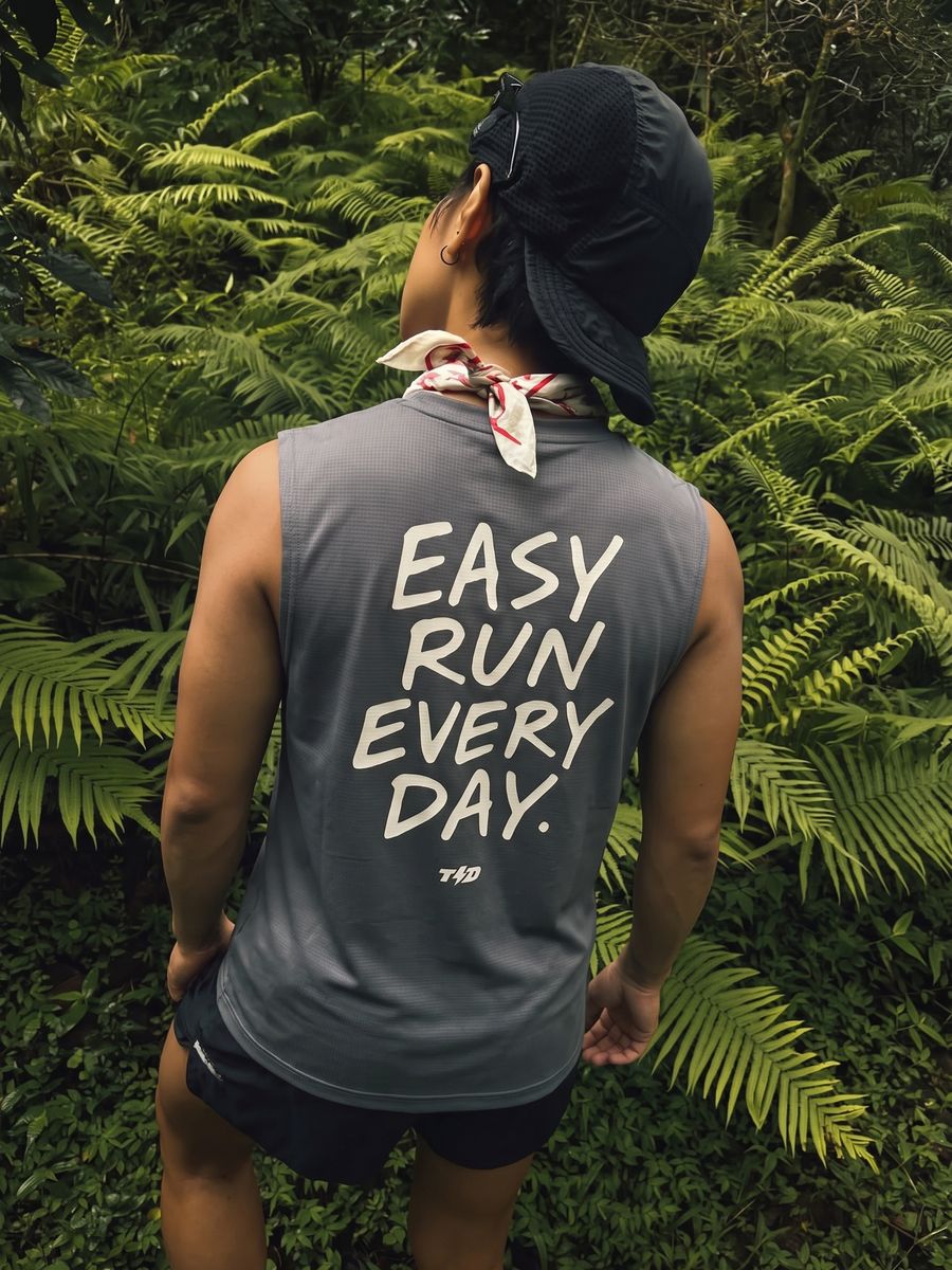 Man in a grey sleeveless running tee, seen from behind on a jungle trail at dusk.